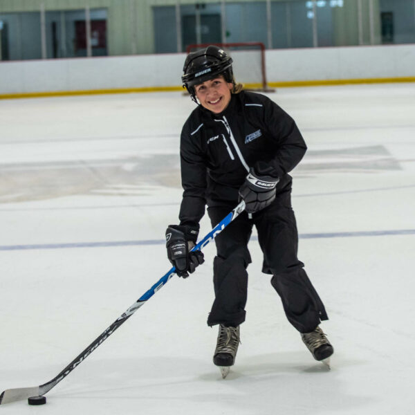 Amy Walsh wearing hockey gear, holding hockey stick in indoor rink.